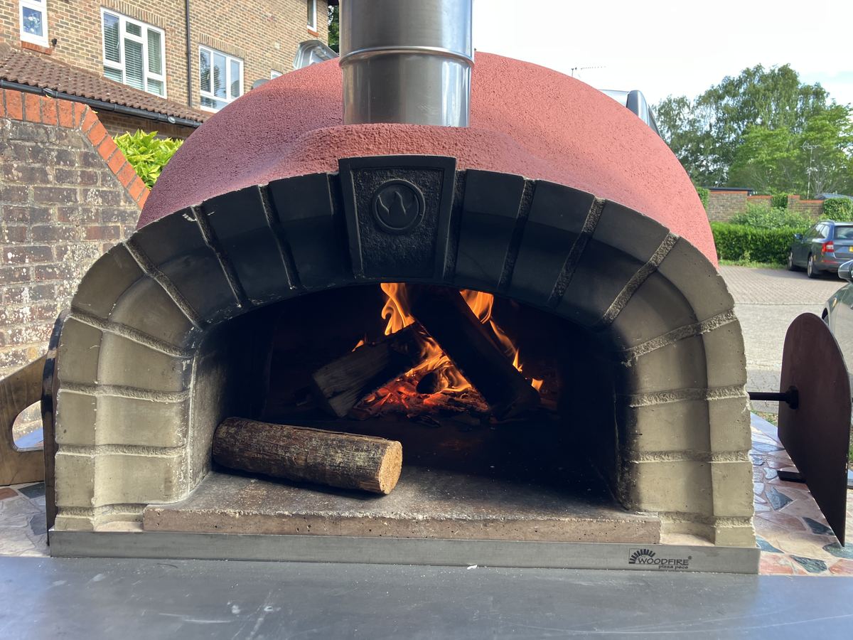 Red dome pizza oven with a lit fire and stacked logs